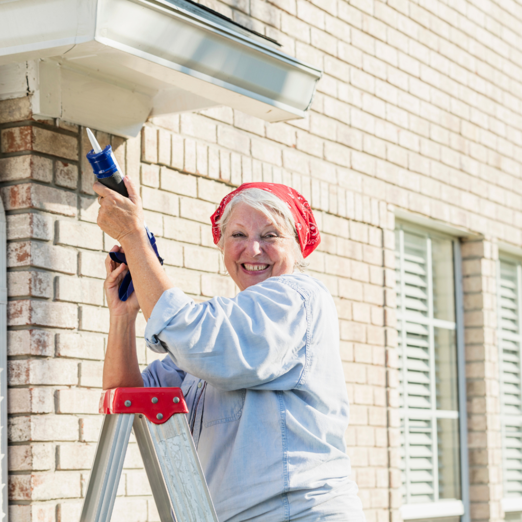 Smiling older woman with a red bandana performing outdoor maintenance using a caulking gun.