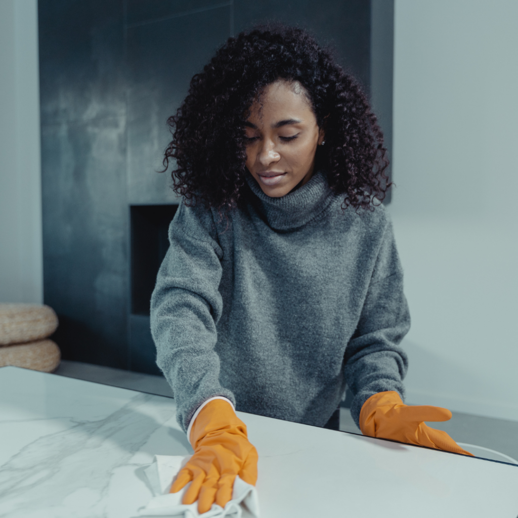 A woman in a gray sweater cleaning a marble countertop with orange gloves.